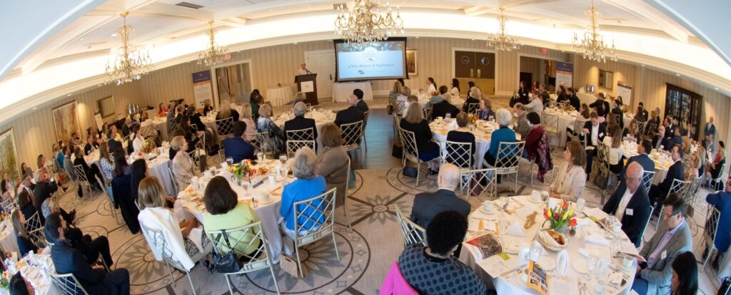 Wide shot of large group of people in banquet hall; at round tables, looking at presentation screen.