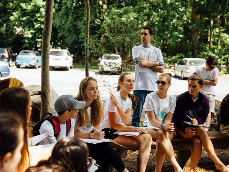 Group of teenagers seated outside on benches, speaking with each other. Parking lot and woods in background.