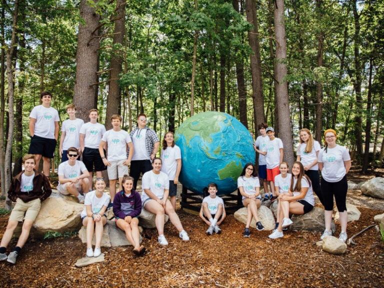 Group of teens in the woods smiling for photo, surrounding a large painted globe.