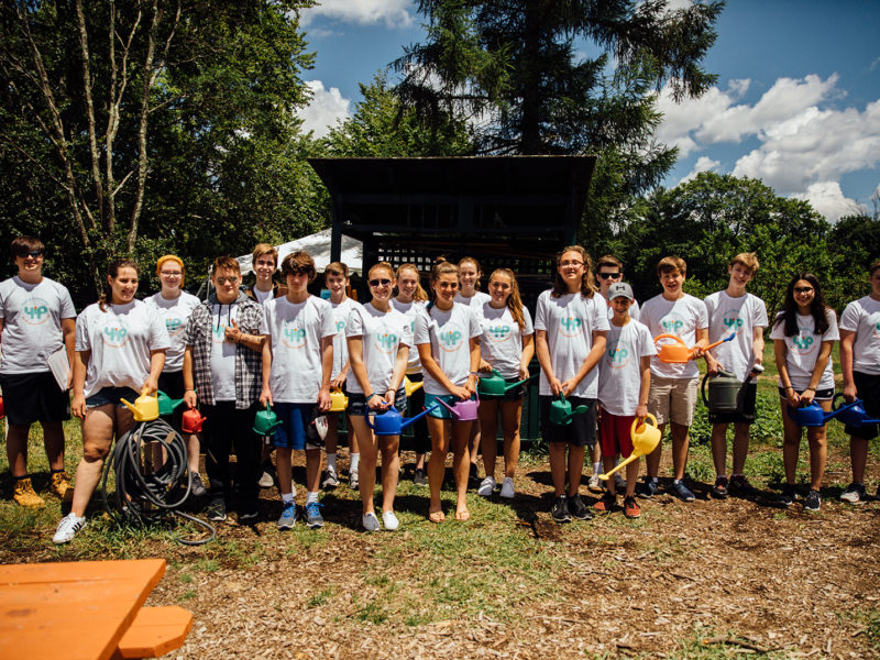Large group of teenagers standing outside with matching white t-shirts, smiling holding watering cans.