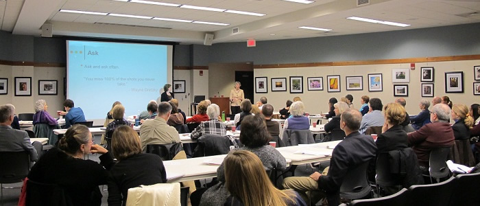 Large room with large group of people watching a presentation screen at front. 2 people stand in front.