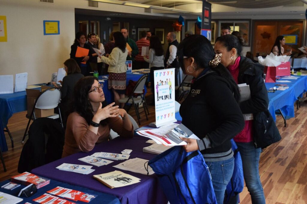 Large room with crowd of people and tables with tablecloths, posters, and flyers. 2 people speak to woman seated at table.