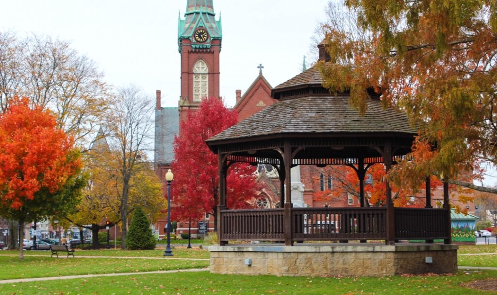 Grass courtyard with dark wooden gazebo at center, and clock tower in background. Red and orange trees in background.