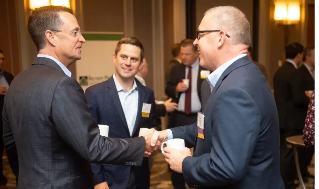 2 people in suits shaking hands, group of people in background.