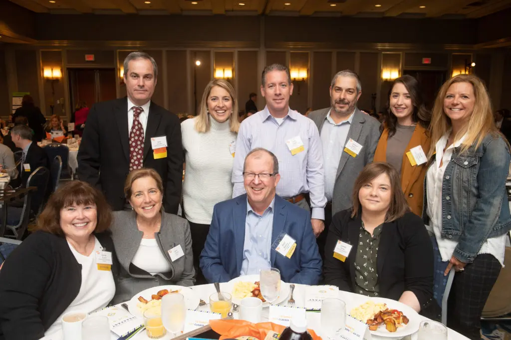 Group of 10 people standing and sitting at round table, in business dress with name tags, smiling for photo.