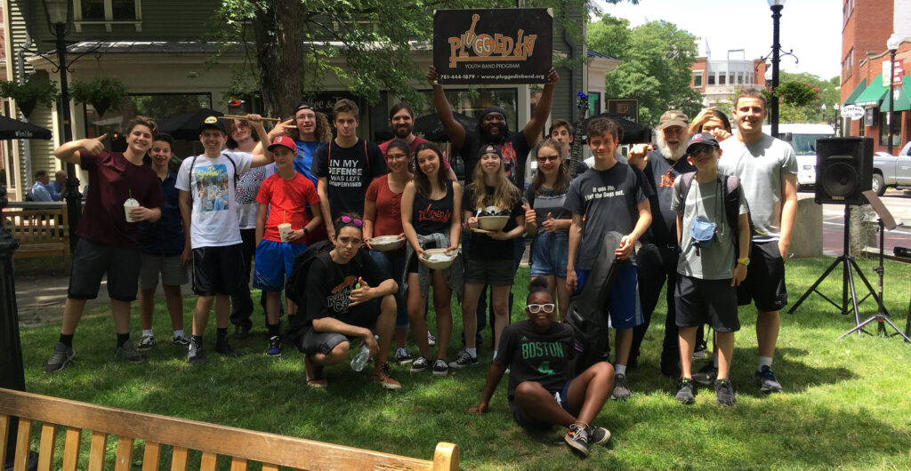 Group of teenagers outside on sunny day smiling in front of sign that says "Plugged In".