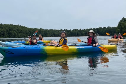 Several kayaks with 2 people in each kayak on the water. People pose with oars and smile for photo. Green forest of trees in background.