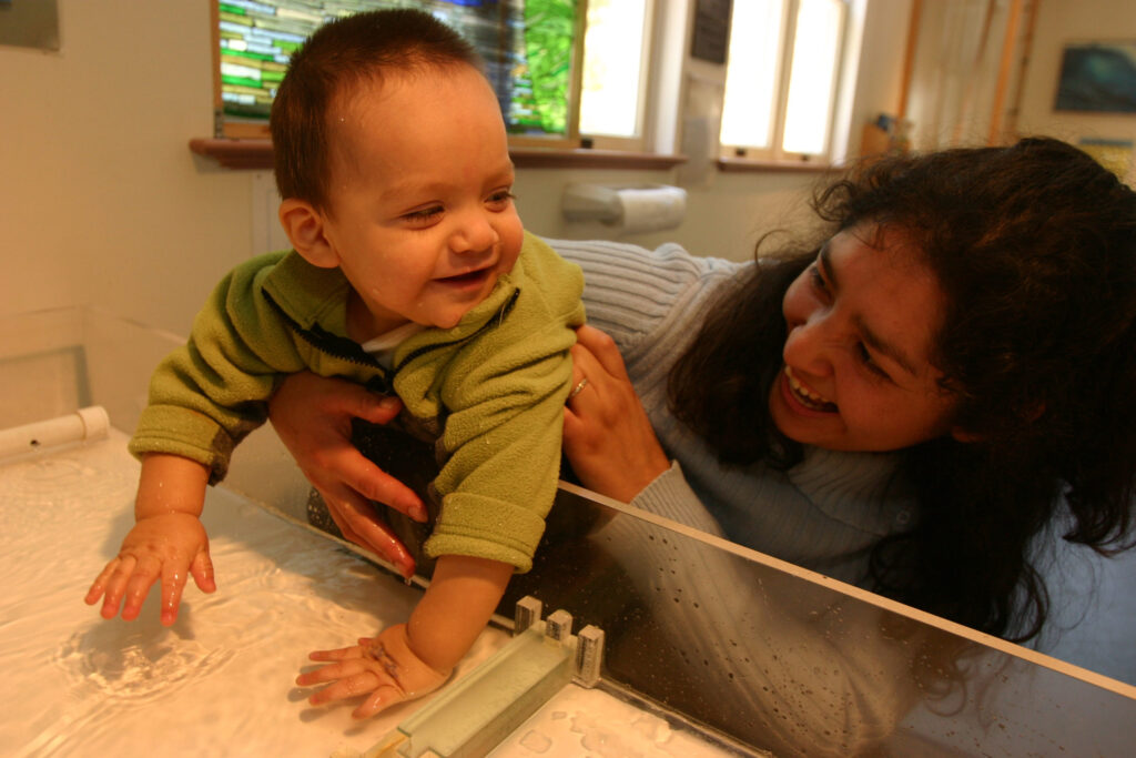 Woman holding toddler, who is smiling and playing in container of water.