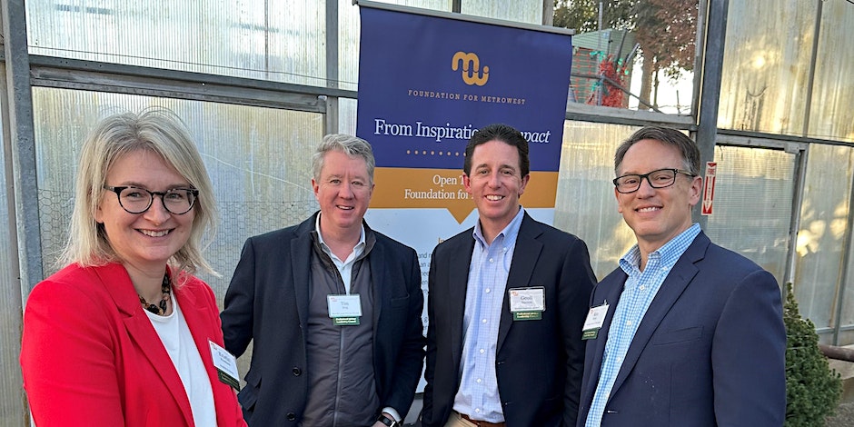 4 people in business dress smile in a group for photo; in front of sign that reads "From Inspiration to Impact"
