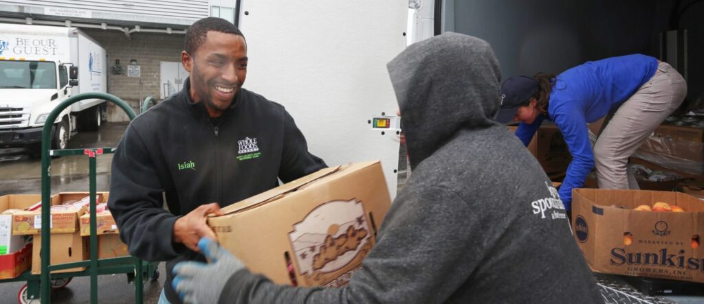 Two people exchanging a cardboard box, a truck is open behind them with boxes of fruit and more people.